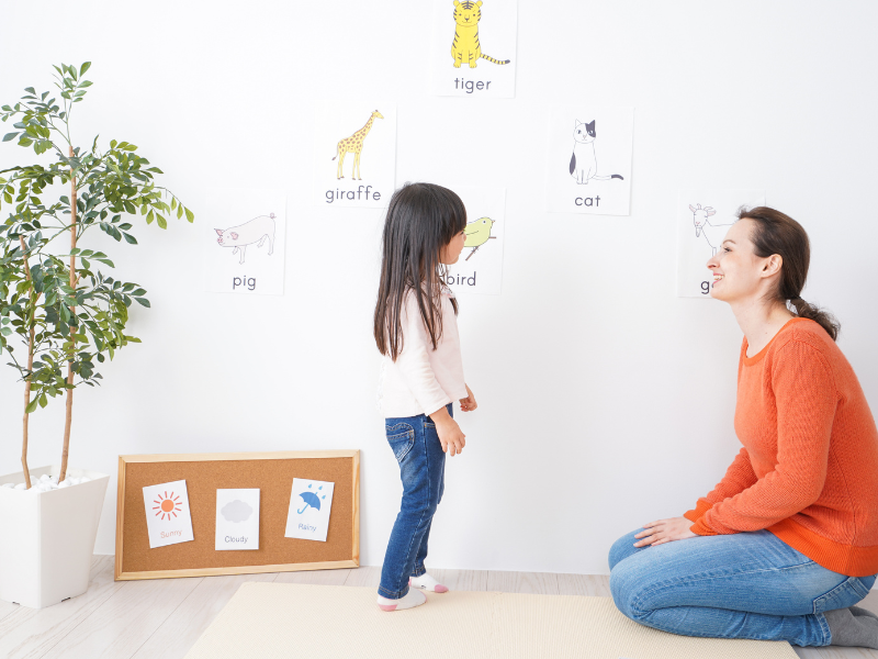 A teacher kneels on the floor, smiling at a young girl who is standing and looking at a wall filled with educational flashcards