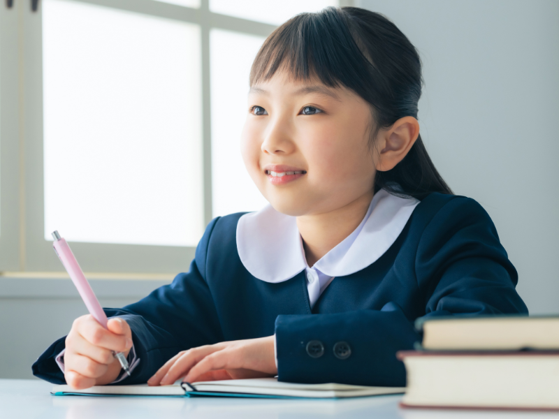 A young girl in a school uniform smiles while writing in a notebook, sitting at a desk with books nearby