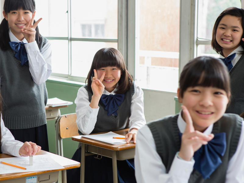 A group of cheerful schoolgirls in uniforms pose for a photo in a classroom, with some making peace signs.